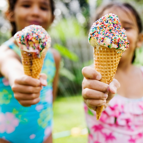 Brightly-colored two ice cream cones with sprinkles being offered with outstretched hands by little girls in bathing suits